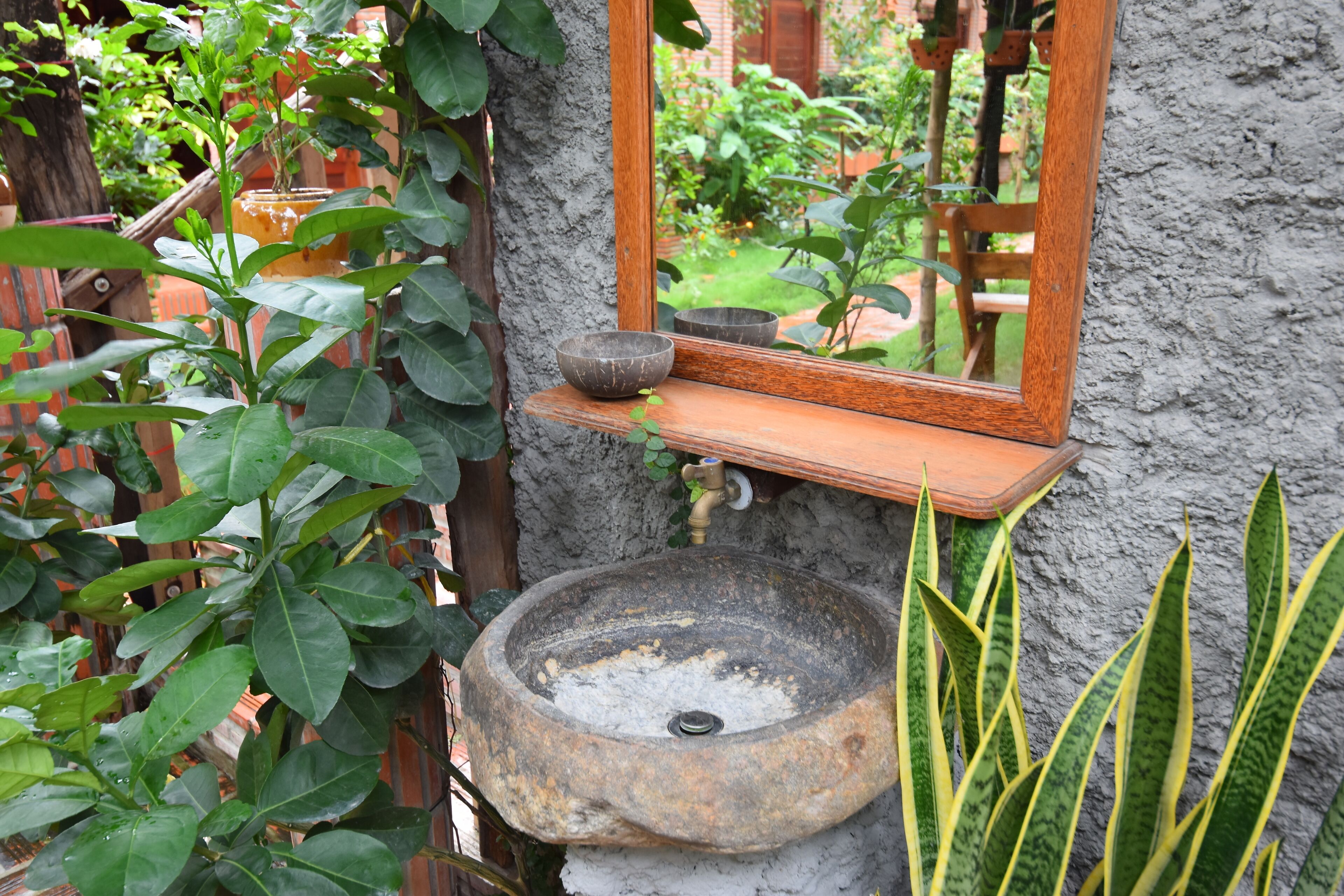 A rustic outdoor setting with a stone basin, a wooden shelf, and a mirror reflecting a garden.