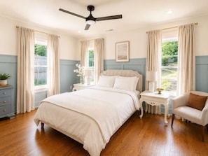 Quarto com cama, poltrona, mesa de cabeceira, luminária, ventilador de teto e janelas com cortinas.