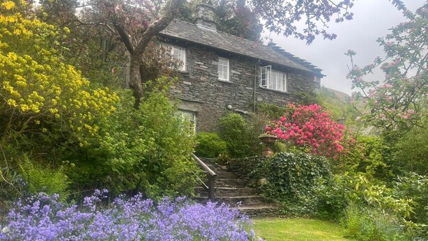 Una casa de piedra con un jardín que incluye flores moradas, un árbol floreciente de color rosa y un árbol floreciente amarillo.