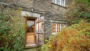 Un edificio de piedra con puerta y ventana de madera, rodeado de follaje denso y un sendero de piedra.