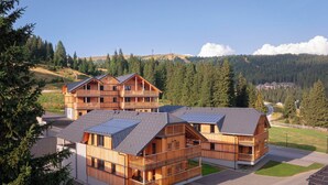 A modern wooden lodge with multiple balconies and solar panels, surrounded by a forested landscape.