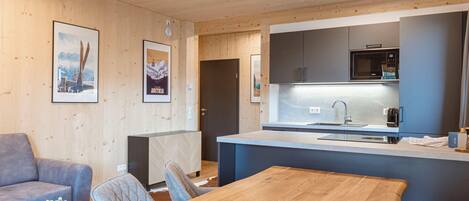 Eine moderne Küche mit Mikrowelle, Spüle und Kaffeemaschine. Im Wohnbereich befindet sich ein Holztisch mit Stühlen sowie eine Couch.