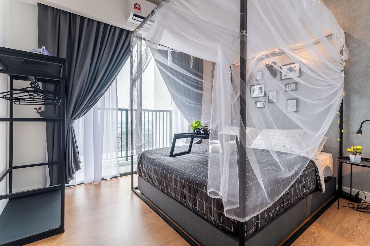 Un dormitorio moderno con una cama bajo un dosel, un estante metálico negro con ropa, una mesita con una planta y una pared adornada con cuadros.