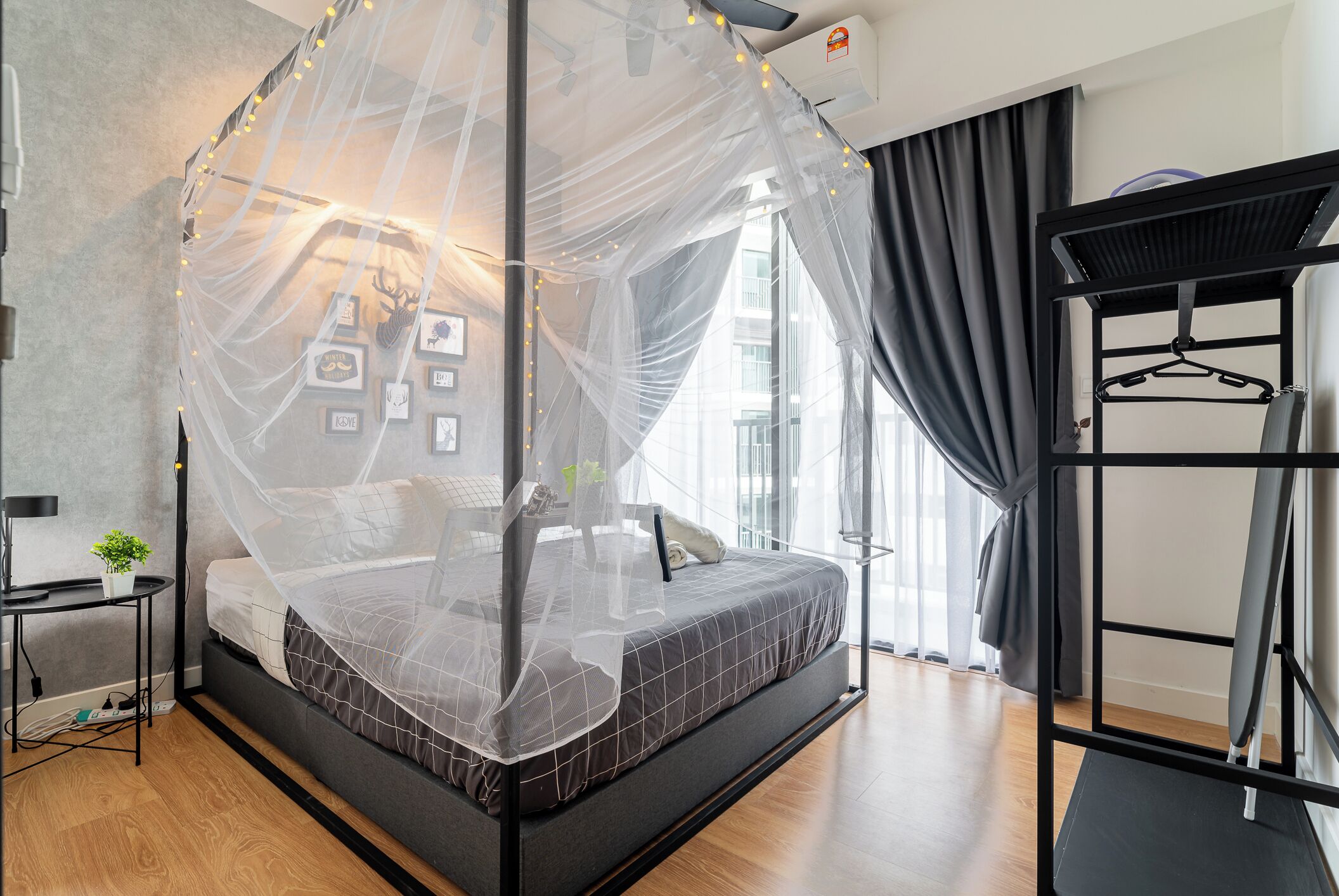 A modern bedroom with a bed under a mosquito net, a bedside table with a plant, and a black metal wardrobe.
