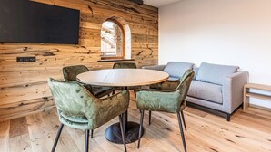 Un salon moderne avec un mur en bois, une table ronde en bois, des fauteuils en velours vert, un canapé gris et un téléviseur à écran plat fixé au mur.