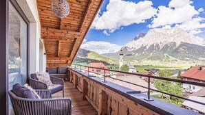 Un balcón de madera con sillas de mimbre y una lámpara colgante, con vistas a un paisaje montañoso que incluye una iglesia.