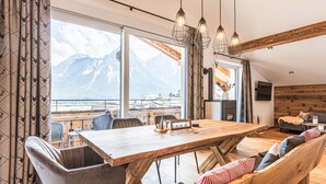 Un comedor amplio con una mesa y sillas de madera, ventanas grandes con vistas a la montaña y un rincón de descanso acogedor con cojines.