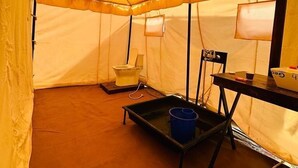 Una carpa con un inodoro, una silla, una mesa y un cubo azul.