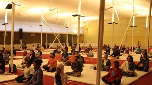 Un grupo de personas sentadas en esterillas en una sala amplia con ventiladores de techo.
