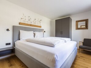 A modern bedroom with a large bed, grey headboard, and a wooden shelf above the bed. There is a grey wardrobe and a chair in the room. - mit 3 Schlafzimmern für 6 Personen by Interhome (Lermoos)