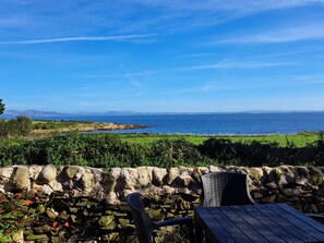 A stone wall with a wicker chair and a wooden table, overlooking a coastal landscape with a view of the sea and distant hills.