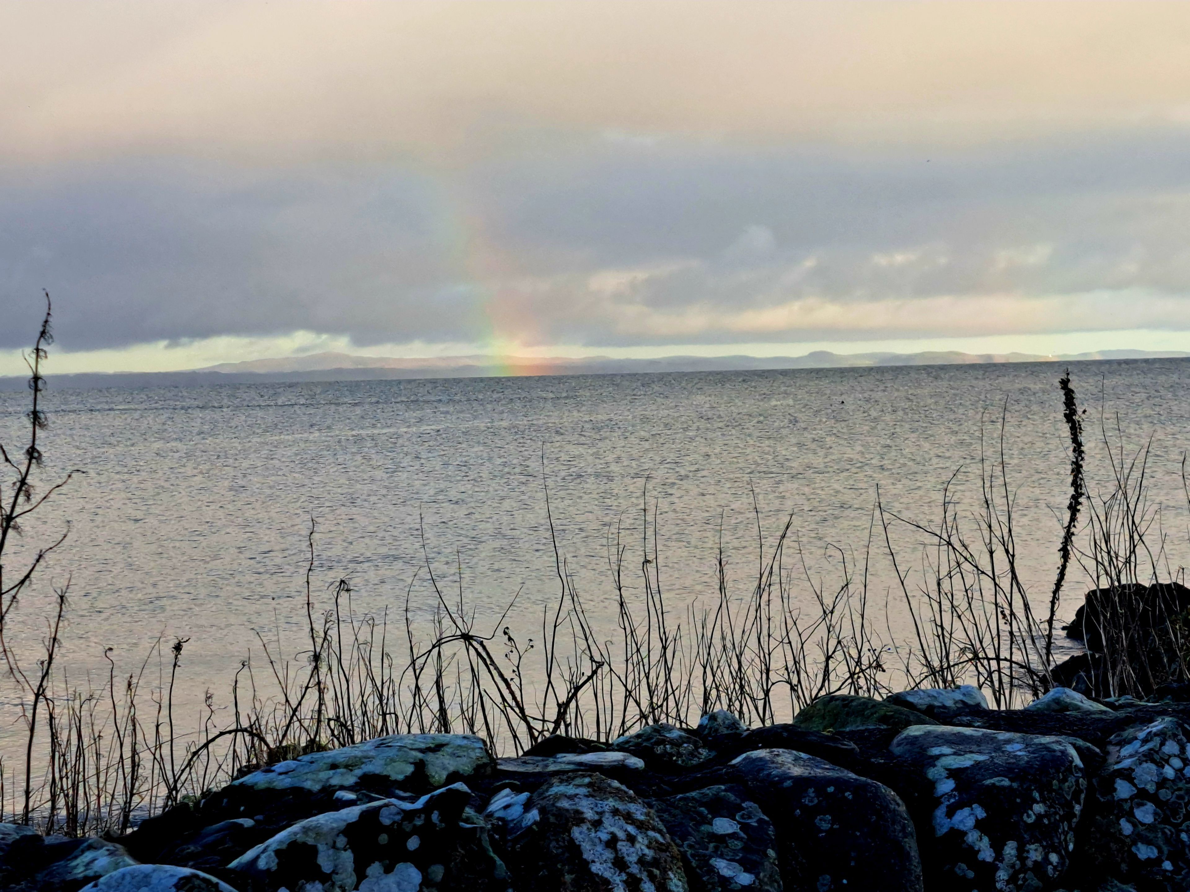 A body of water with a rainbow, rocky shoreline, and distant mountains.