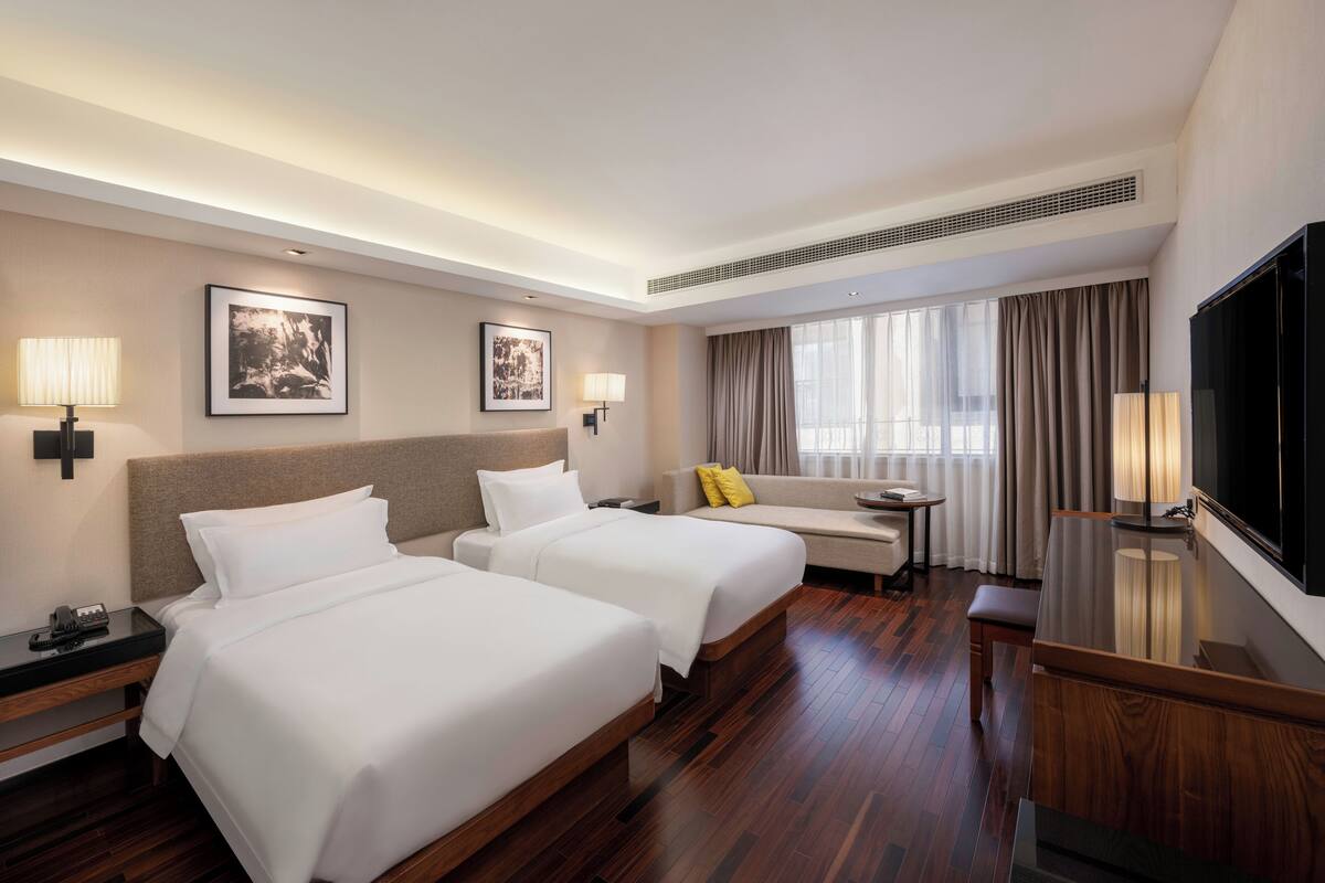 A hotel room with two beds, a TV, a desk, and a window with curtains.
