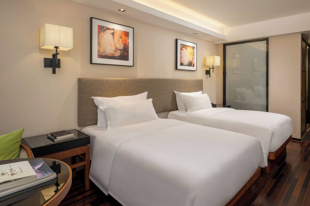 A hotel room with two single beds, a nightstand with a lamp, a small table with a green cushion, and framed artwork on the walls.