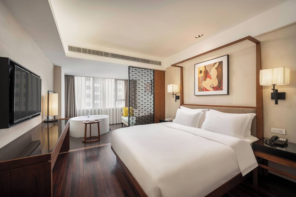 A hotel room with a large bed, a flat-screen TV, a desk with a lamp, a bathroom with a shower, and a window with curtains.