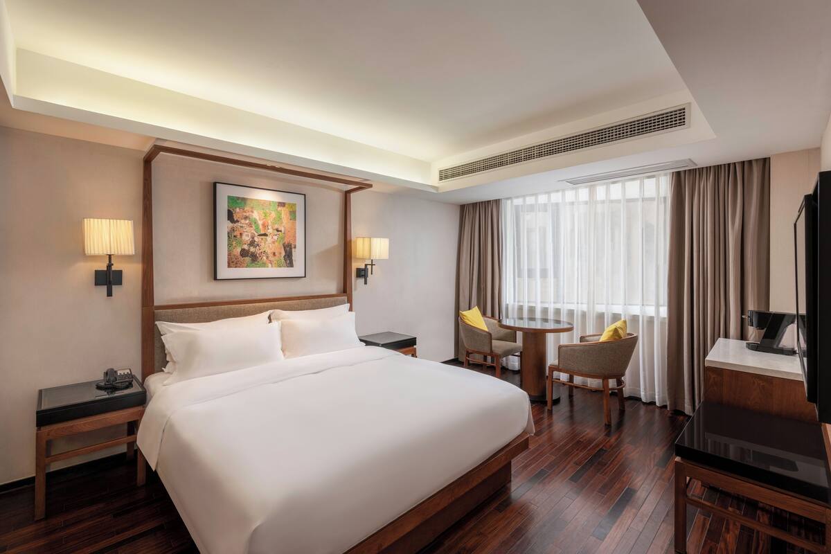 A hotel room with a large bed, two bedside tables, a desk with a chair, a TV, and a large window with curtains.