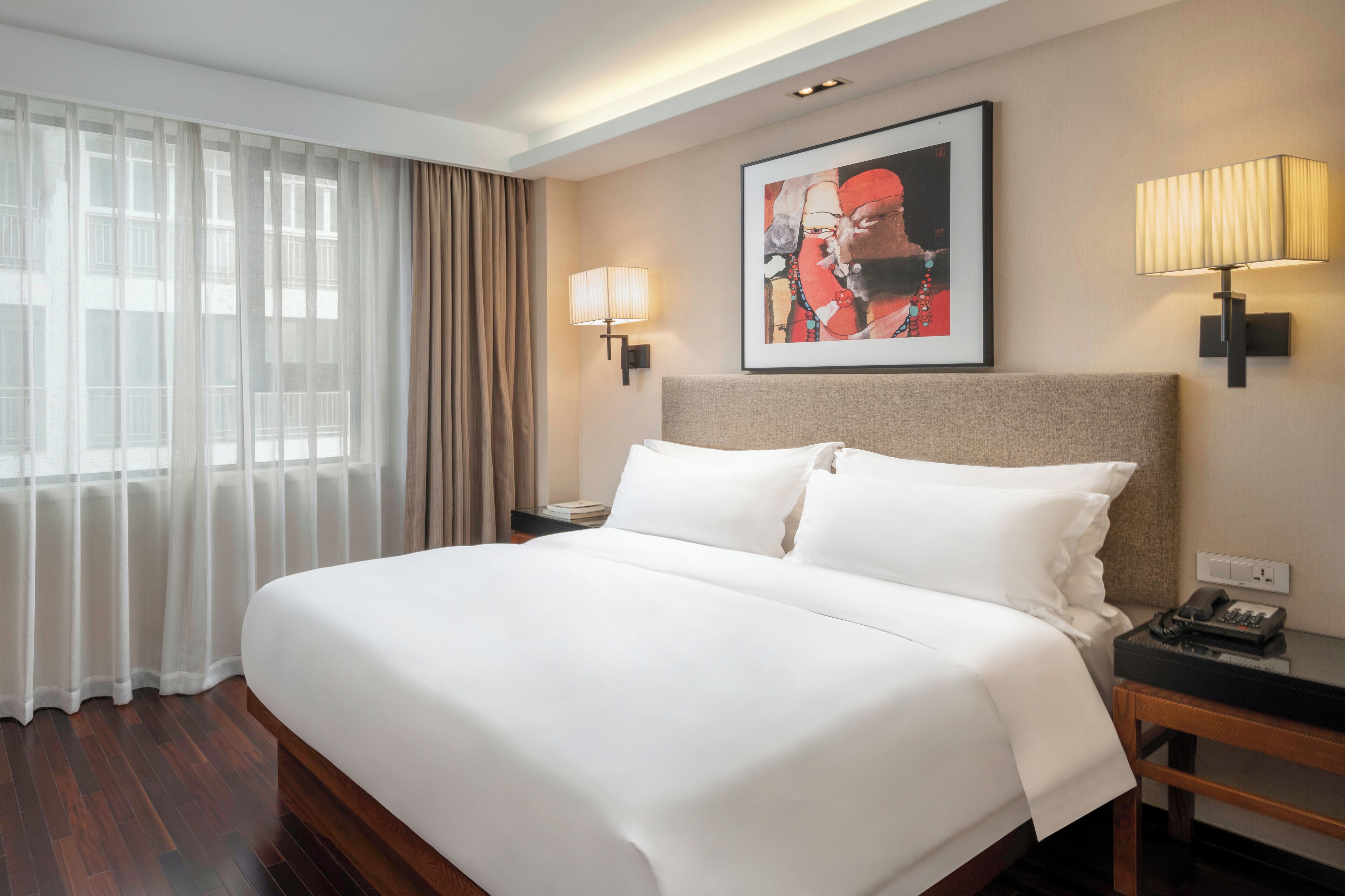 Una habitación de hotel con una cama grande, dos lámparas de noche, un cuadro en la pared y una ventana con cortinas translúcidas.