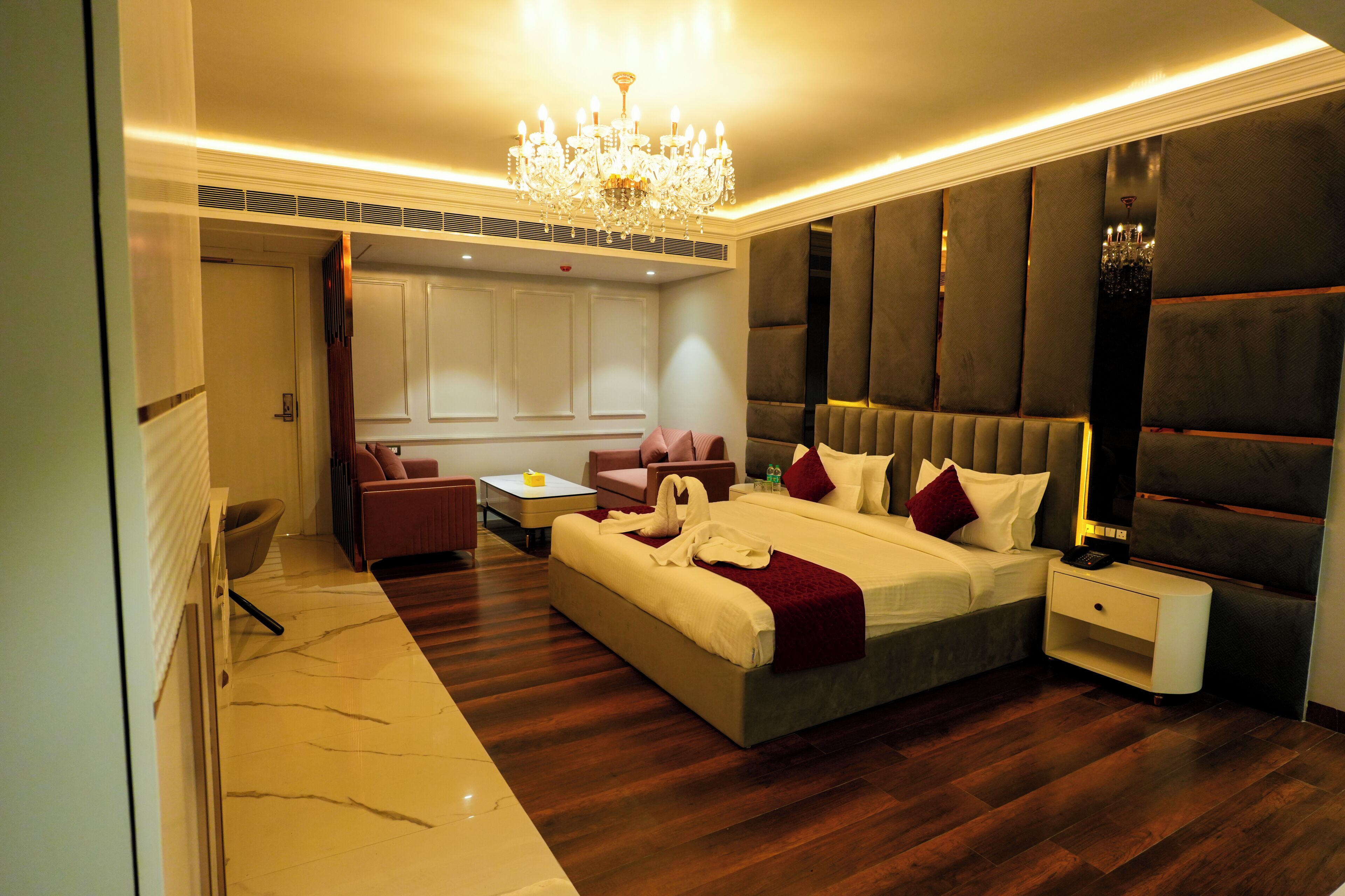 A modern hotel room with a large bed, a sitting area with a sofa and armchairs, a chandelier, and a view of the city.