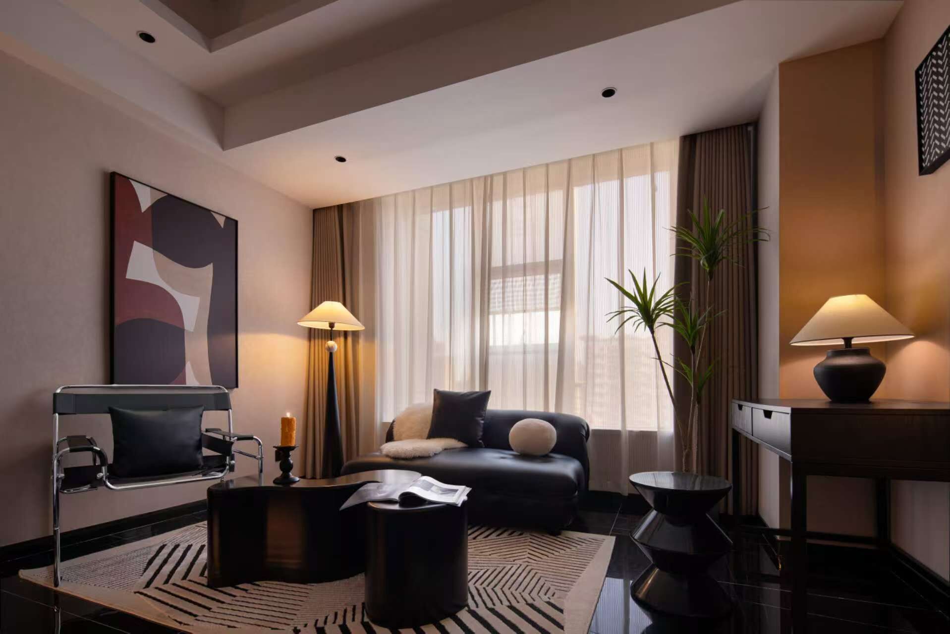 A modern hotel room with a black leather sofa, a black armchair, a round coffee table, a side table with a lamp, and a large window with sheer curtains.