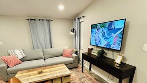 A living room with a grey sofa, a brown leather chair, a wooden coffee table, a patterned rug, a black TV stand with a flat-screen TV, and a grey curtain. - Chic 1-bedroom apartment in  Springtown, TX with AC, WiFi (Springtown)