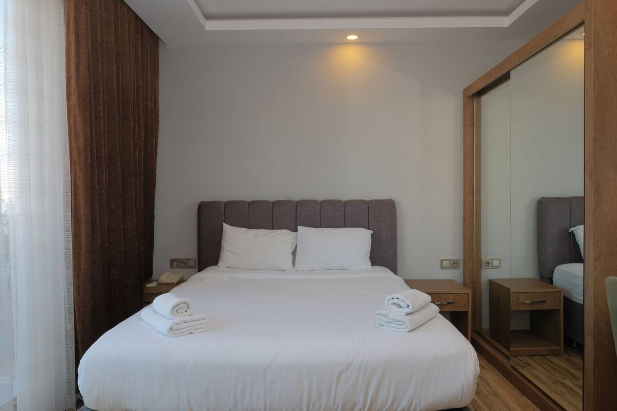 Una cama bien hecha con sábanas blancas y dos toallas enrolladas, una mesita de noche de madera con un teléfono, y un armario con espejo y marco de madera.