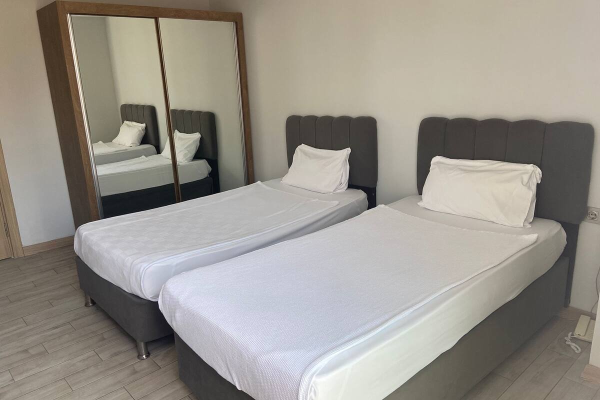 Una cama doble con cabecera gris y ropa de cama blanca, un espejo grande y un armario con estructura de madera.