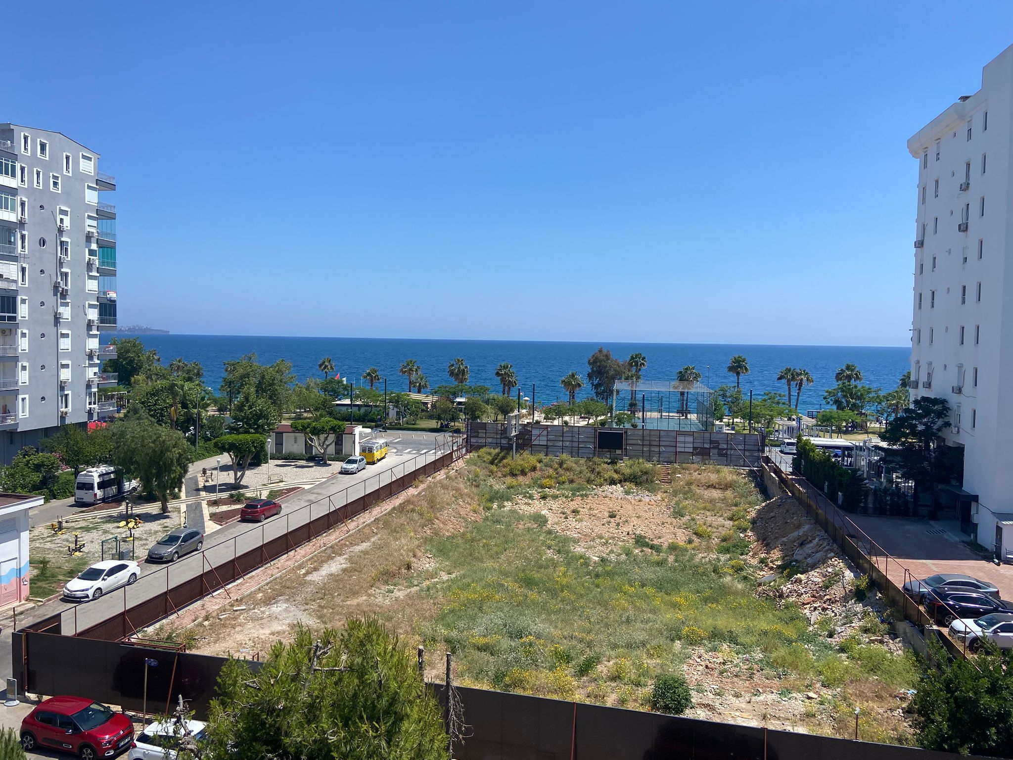 A coastal view with a tennis court, palm trees, and a road with parked cars.
