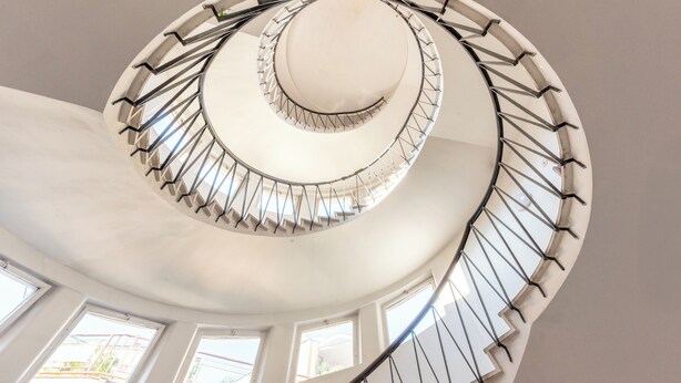 A spiral staircase with a white interior and metal railings, leading to an upper floor with windows.