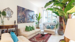 A living room with a sofa set, a coffee table, a large painting, and several potted plants.
