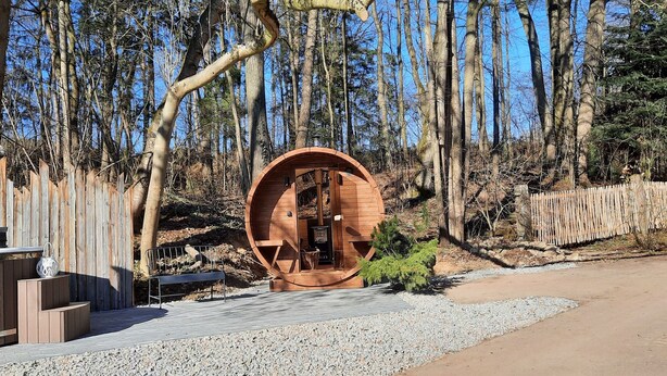 A wooden structure resembling a barrel with a bench and a small table, surrounded by a wooden fence and trees. - Vacation home Rötzer - Forsthausschmankerl (Bruck in der Oberpfalz)