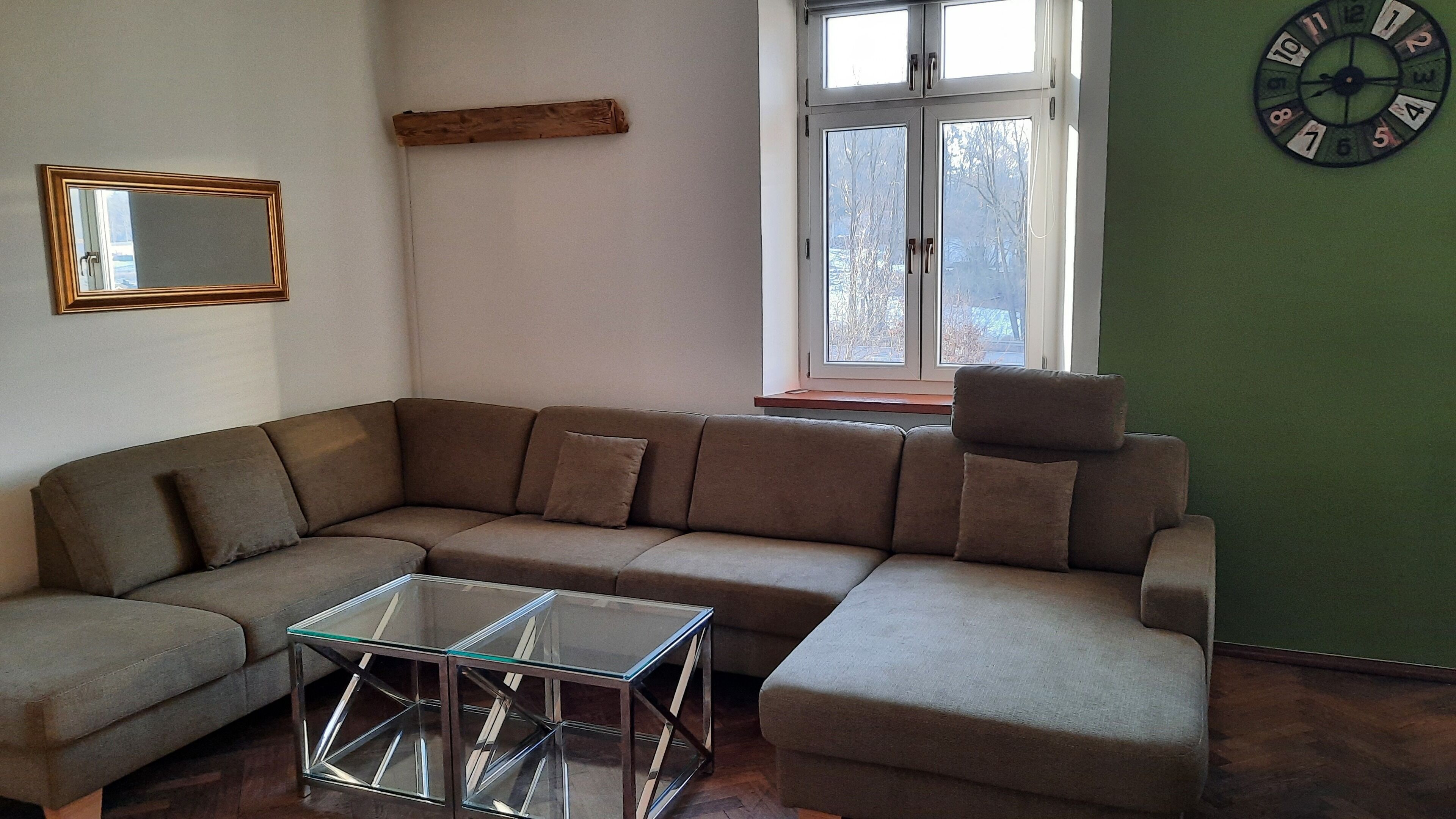 Una sala de estar con un sofá modular, una mesa de centro de vidrio, un estante de madera, una ventana con vistas y un reloj de pared.