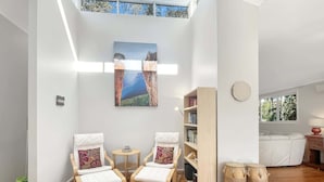 A spacious living area with a high ceiling, wooden flooring, two white armchairs with patterned cushions, a small round table, a bookshelf, and a large painting above the chairs. - Riverside 3-Bed Townhouse with Spacious Deck (Mossy Point)