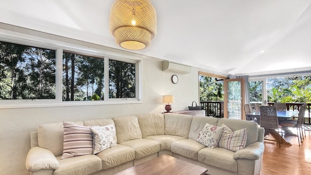 A living room with a large L-shaped sofa, a wooden coffee table, and a hanging light fixture. There are large windows with a view of trees and a balcony with outdoor seating. - Riverside 3-Bed Townhouse with Spacious Deck (Mossy Point)