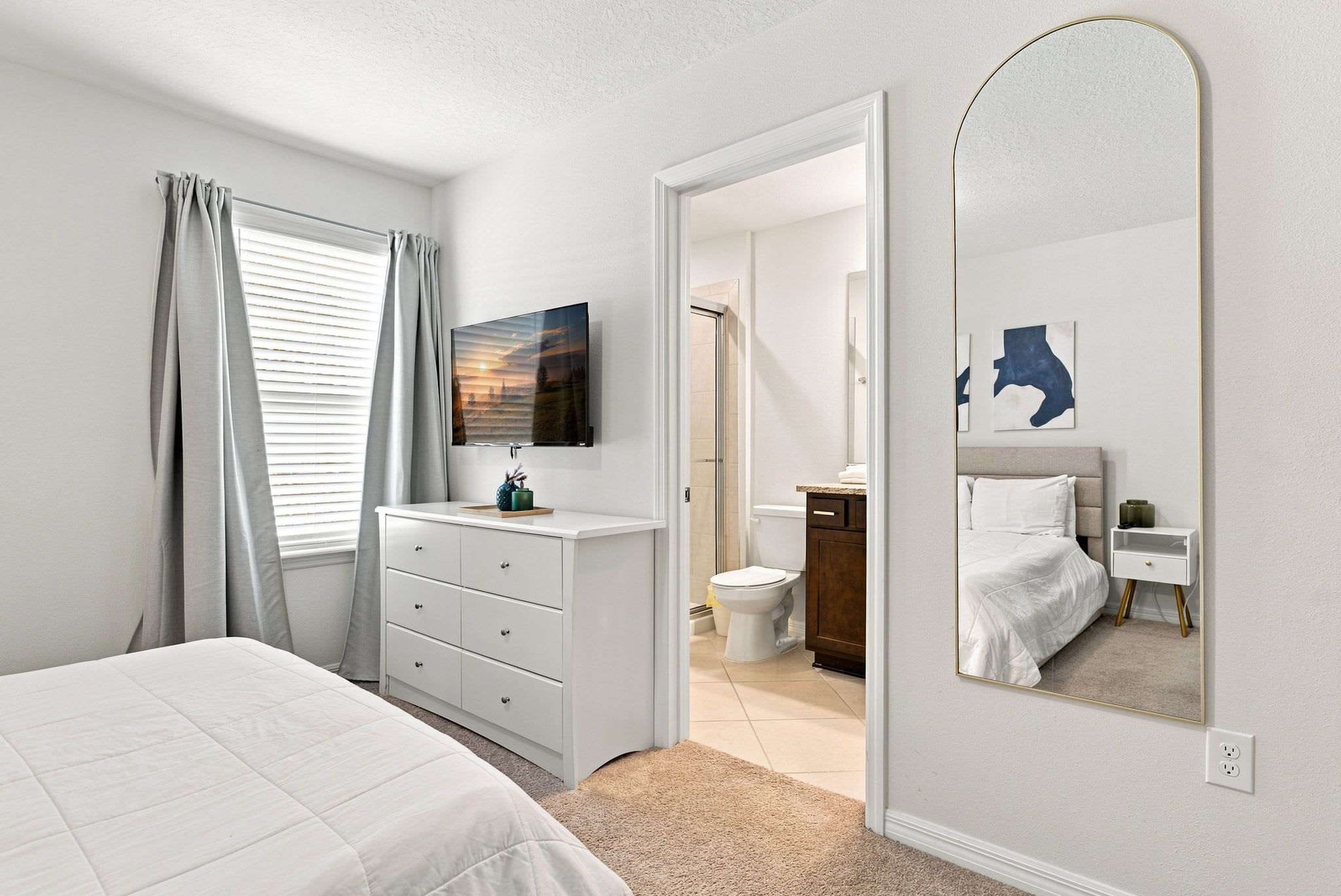 Un dormitorio con cama, cómoda, televisor, espejo y baño visible a través de una puerta abierta.