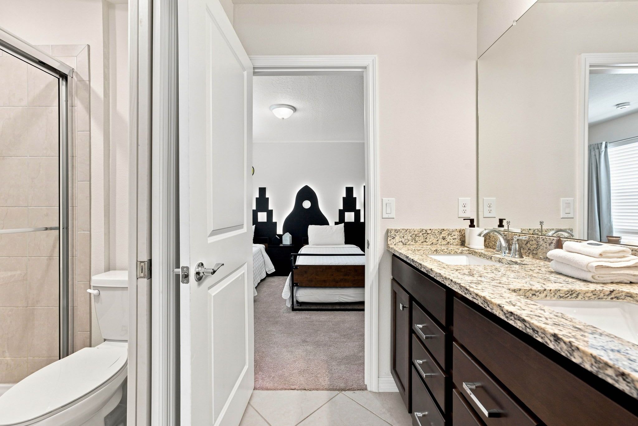 Une salle de bain comprenant des toilettes, un lavabo et un miroir. Une porte donne accès à une chambre à coucher équipée d’un lit et d’un luminaire fixé au mur.