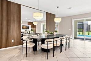 A modern dining area with a round table, multiple chairs, pendant lighting, and a view of a pool through large windows. - Stunning 10 Bed Home with Themed Rooms, Pool (Clermont)