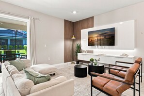 Ein modernes Wohnzimmer mit einer Couch, Sesseln, einem Couchtisch, einem an der Wand befestigten Fernseher und Blick ins Freie durch große Fenster.