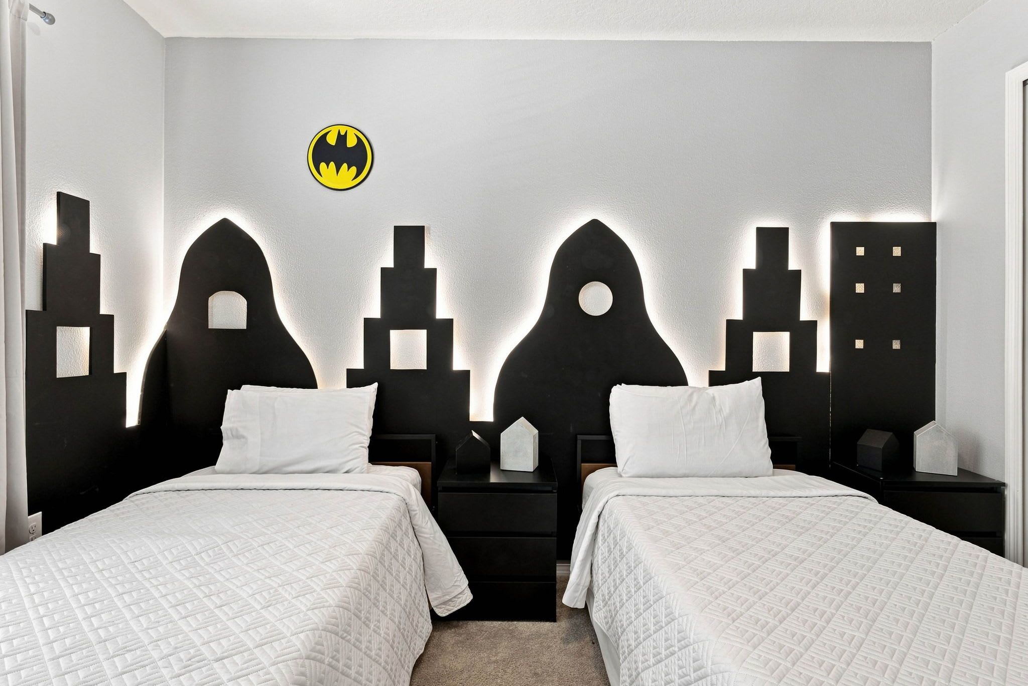 A bedroom with two beds, a wall-mounted Batman logo, and a cityscape design on the wall.