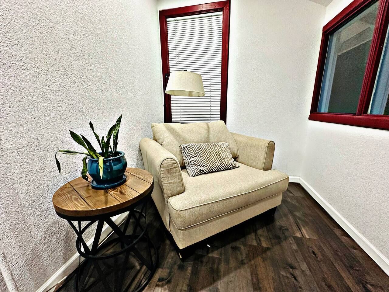 A corner room with a beige sofa, a small round table with a blue pot, a lamp, and a window with blinds.