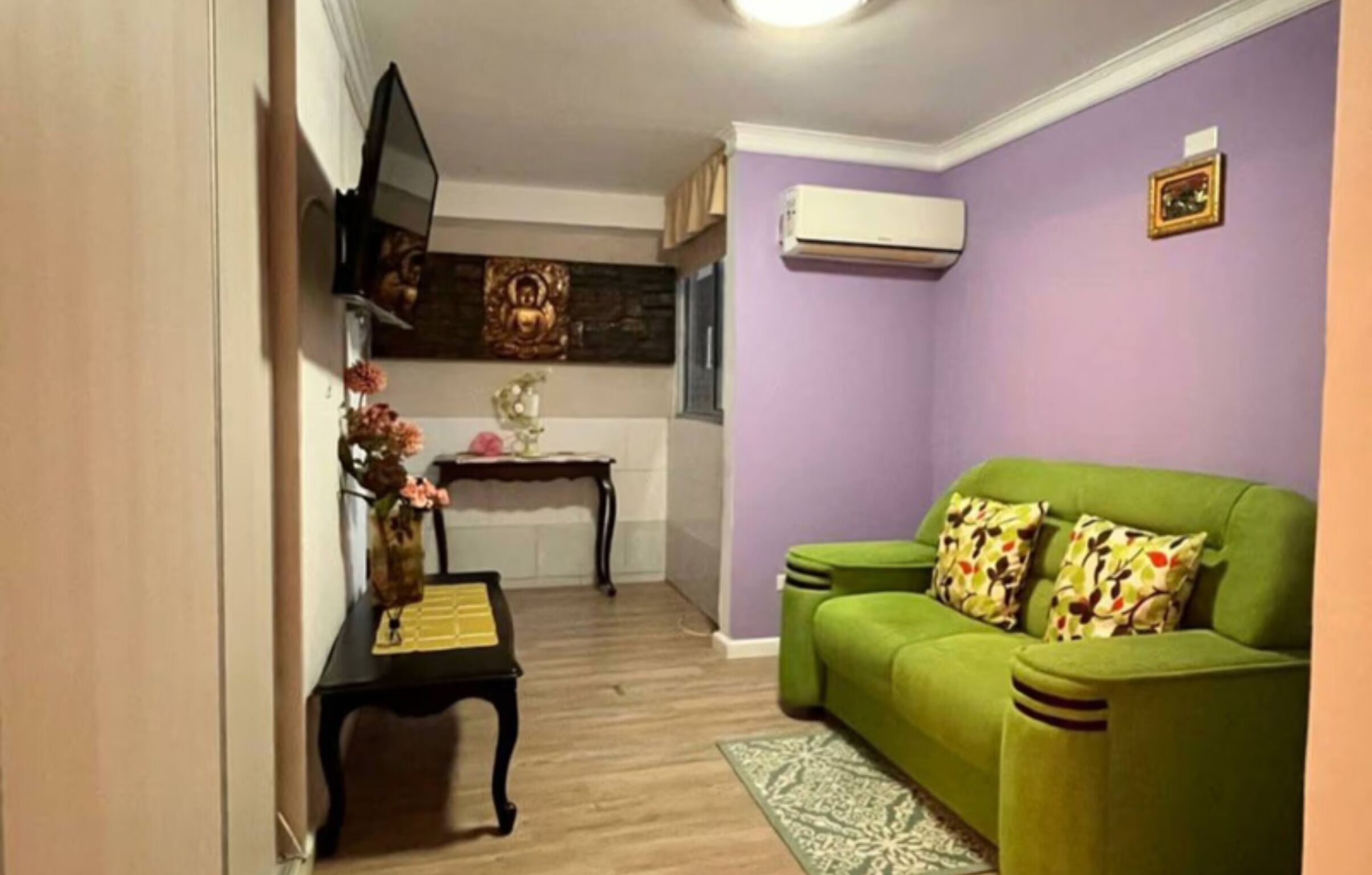 A living room with a green sofa, a small table with a vase of flowers, and a wall-mounted TV.