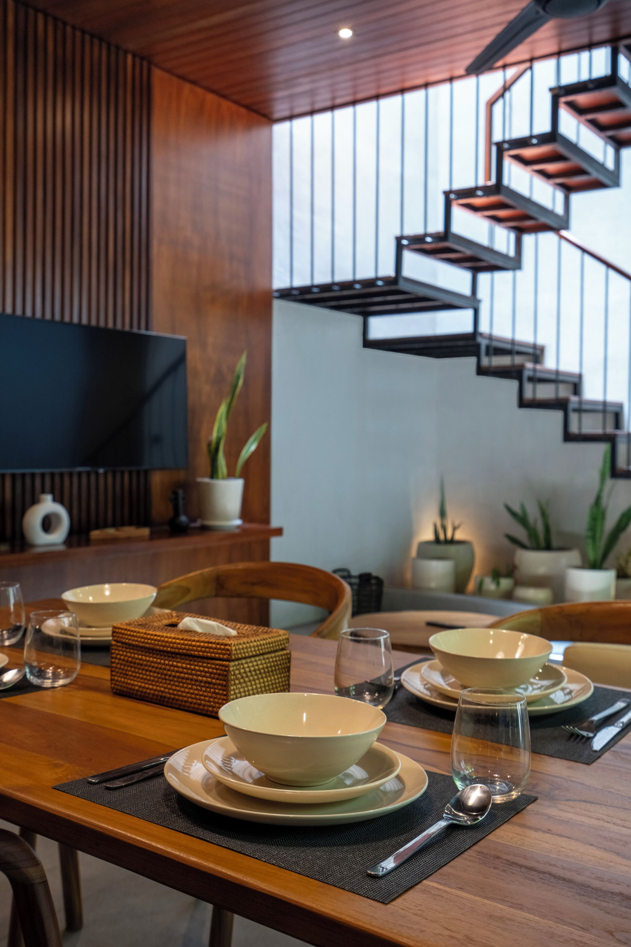 A dining area with a wooden table set for four, a modern staircase, and a flat-screen TV on the wall.