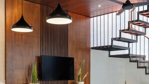 Un comedor moderno con una mesa de madera, sillas e iluminación colgante. Un televisor de pantalla plana está montado en la pared, y hay plantas en macetas sobre la mesa.
