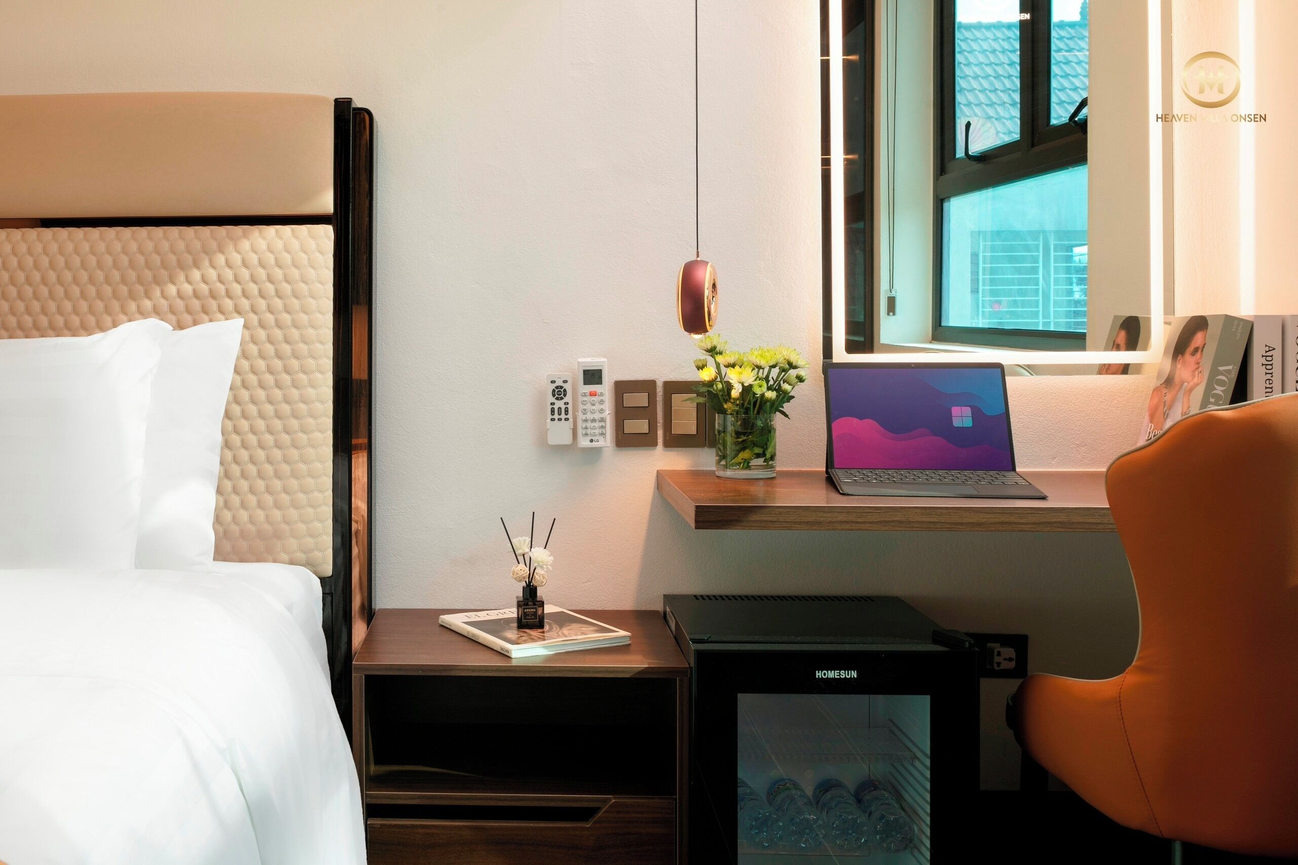 Une chambre d ’hôtel avec un lit, un bureau avec un ordinateur portable, un petit réfrigérateur, une table de chevet avec un livre et un vase de fleurs, et une fenêtre donnant sur des immeubles.