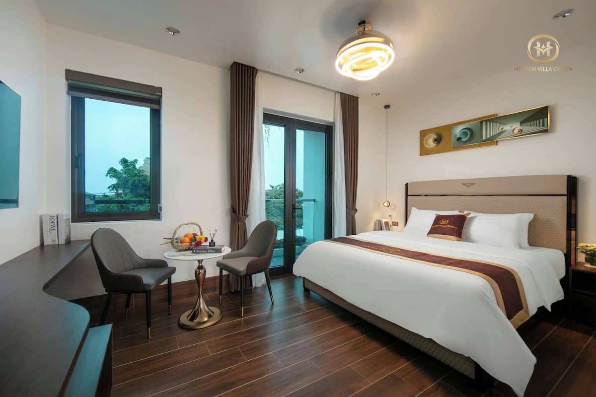 A hotel room with a large bed, a desk with two chairs, a small table with a basket of fruit, and a large window offering a view of the outdoors.