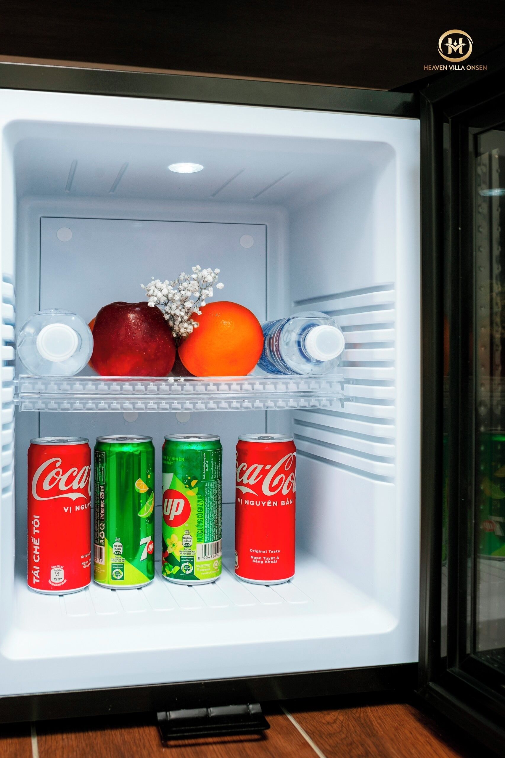 A refrigerator with a clear shelf, containing a bottle of water, an apple, an orange, and three cans of Coca-Cola.