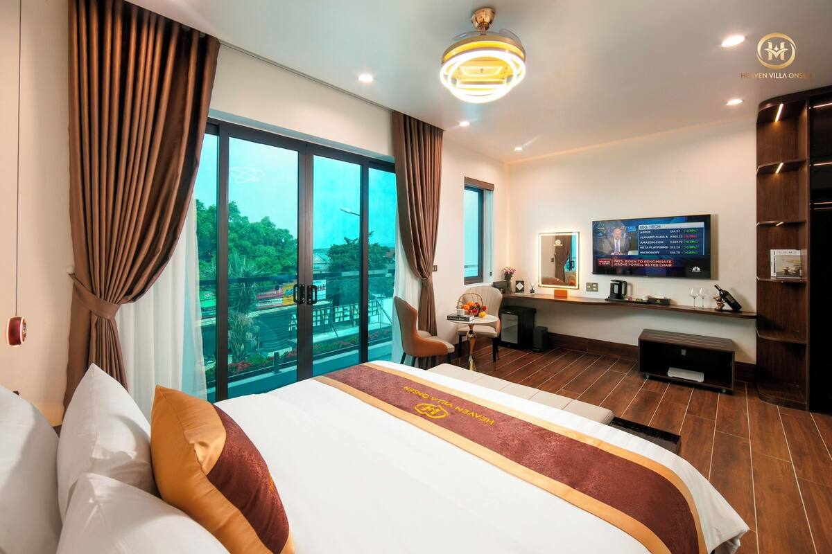 A hotel room with a large bed, a desk, a TV, and a view of greenery outside.