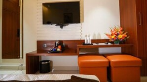 Quarto de hotel com escrivaninha de madeira, televisão na parede, pequena mesa com um vaso de flores e um banco laranja.