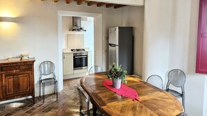 A dining area with a wooden table, clear chairs, a red placemat, and a potted plant. A kitchen with white cabinets and a stainless steel refrigerator is visible in the background. - Manor in Marche With Pool Near Adriatic (Monsano)