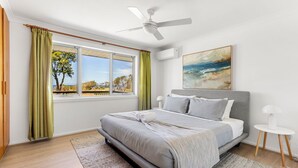 A bedroom with a bed, a ceiling fan, a painting, and a view of the outdoors.
