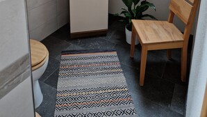 Un baño pequeño y funcional con tragaluz, un mueble de almacenamiento, inodoro, una silla de madera y alfombra estampada.